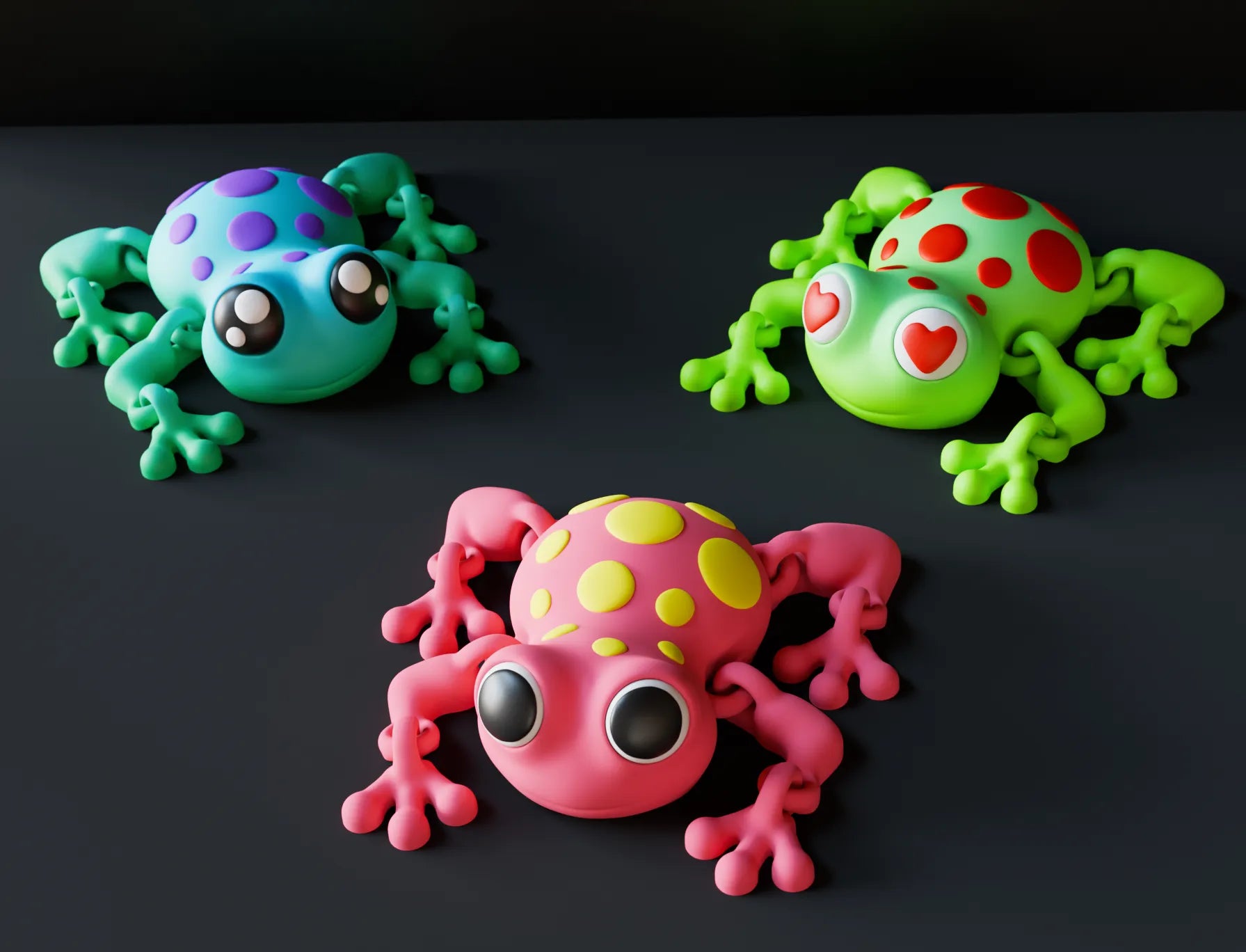 Three brightly colored 3D-printed frog toys with large cartoon eyes, displayed on a black background. One frog is pink with yellow dots, one is teal with purple dots, and one is green with red hearts in its eyes.