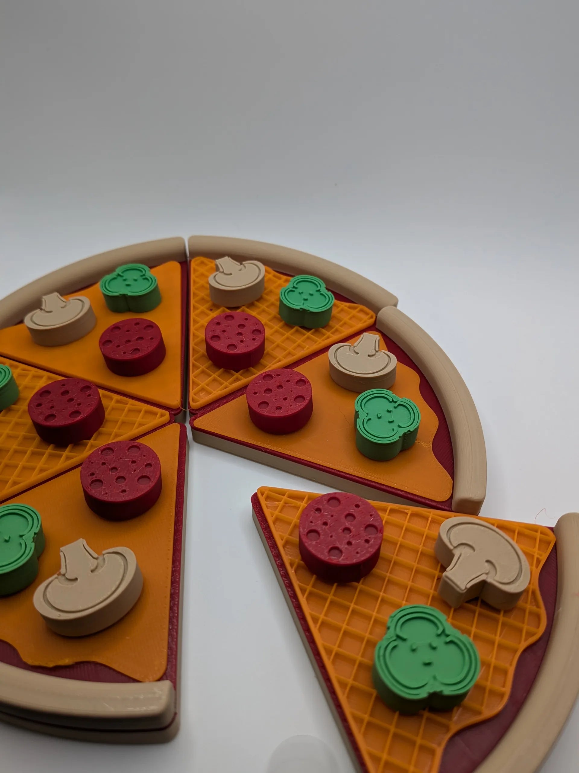 3D-printed toy pizza with six colorful slices, topped with removable pieces like mushrooms, pepperoni, and capsicum, displayed on a white background.