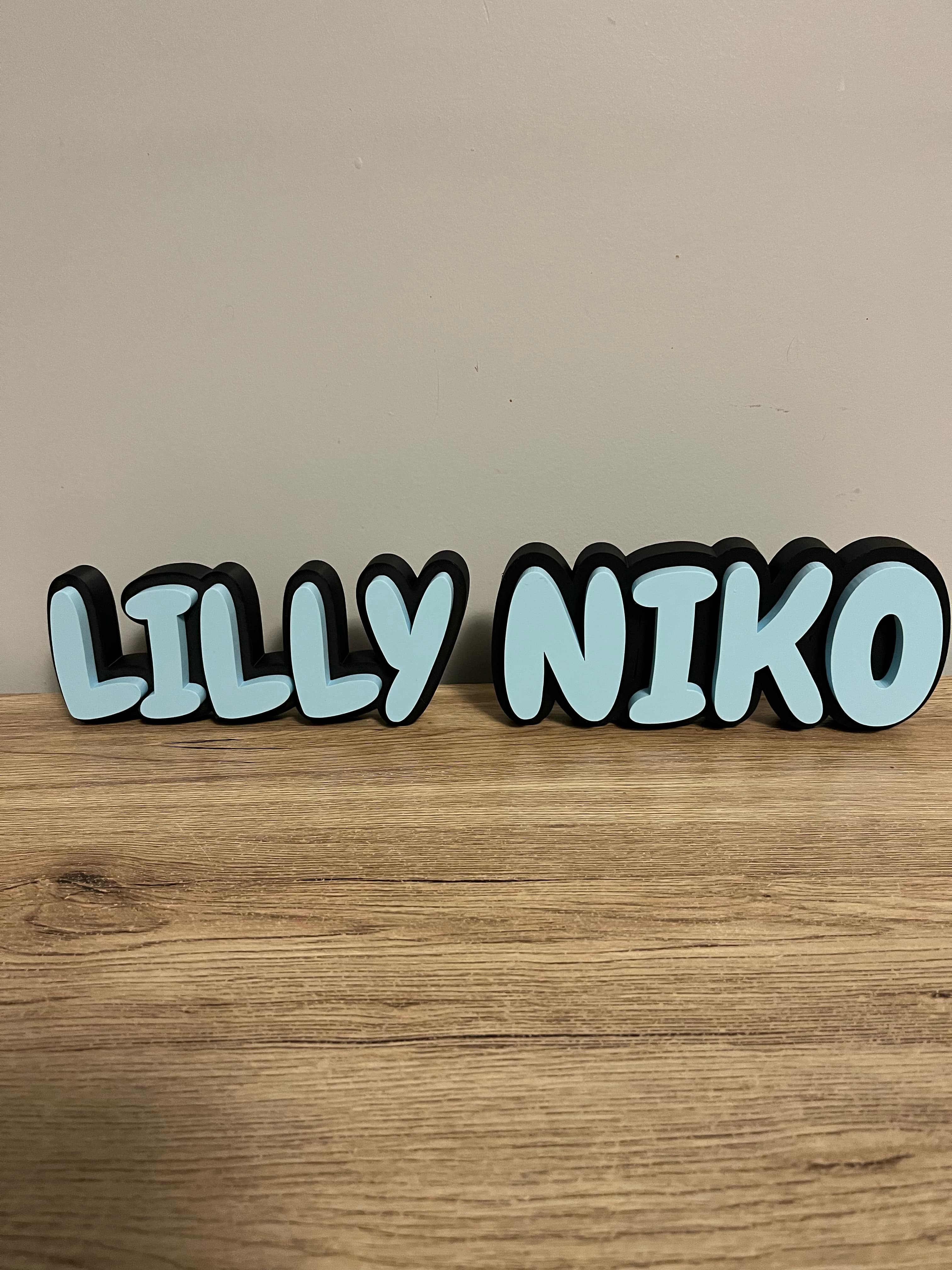 3D-printed name sign in light blue with black outline, reading ‘LILLY NIKO’, displayed on a wooden surface with a neutral wall background.