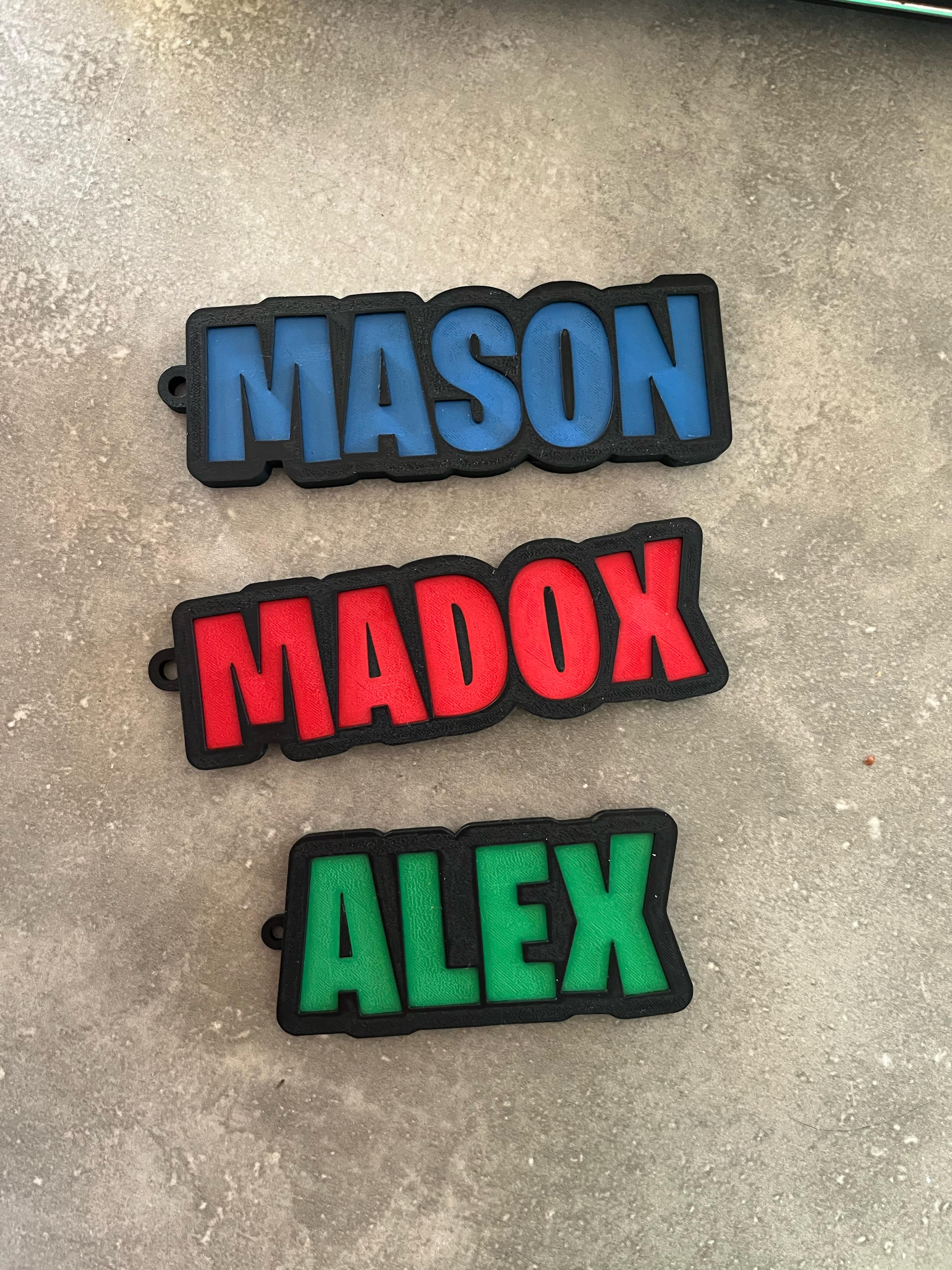 Three colourful 3D-printed name tags reading 'MASON' in blue, 'MADOX' in red, and 'ALEX' in green, each with black borders, displayed on a concrete surface.