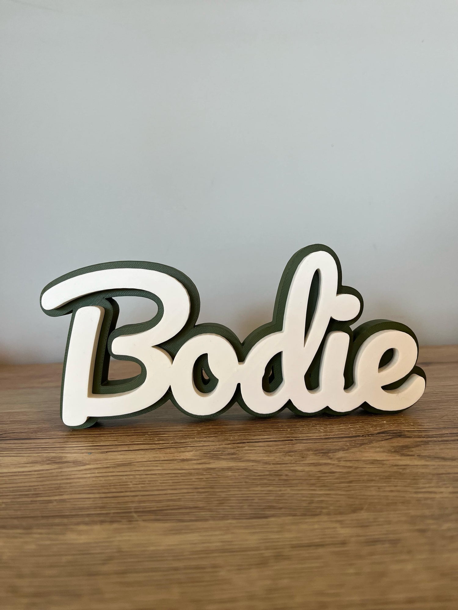 Cream and green 3D-printed name sign reading ‘Bodie’, displayed on a wooden surface with a light grey background.