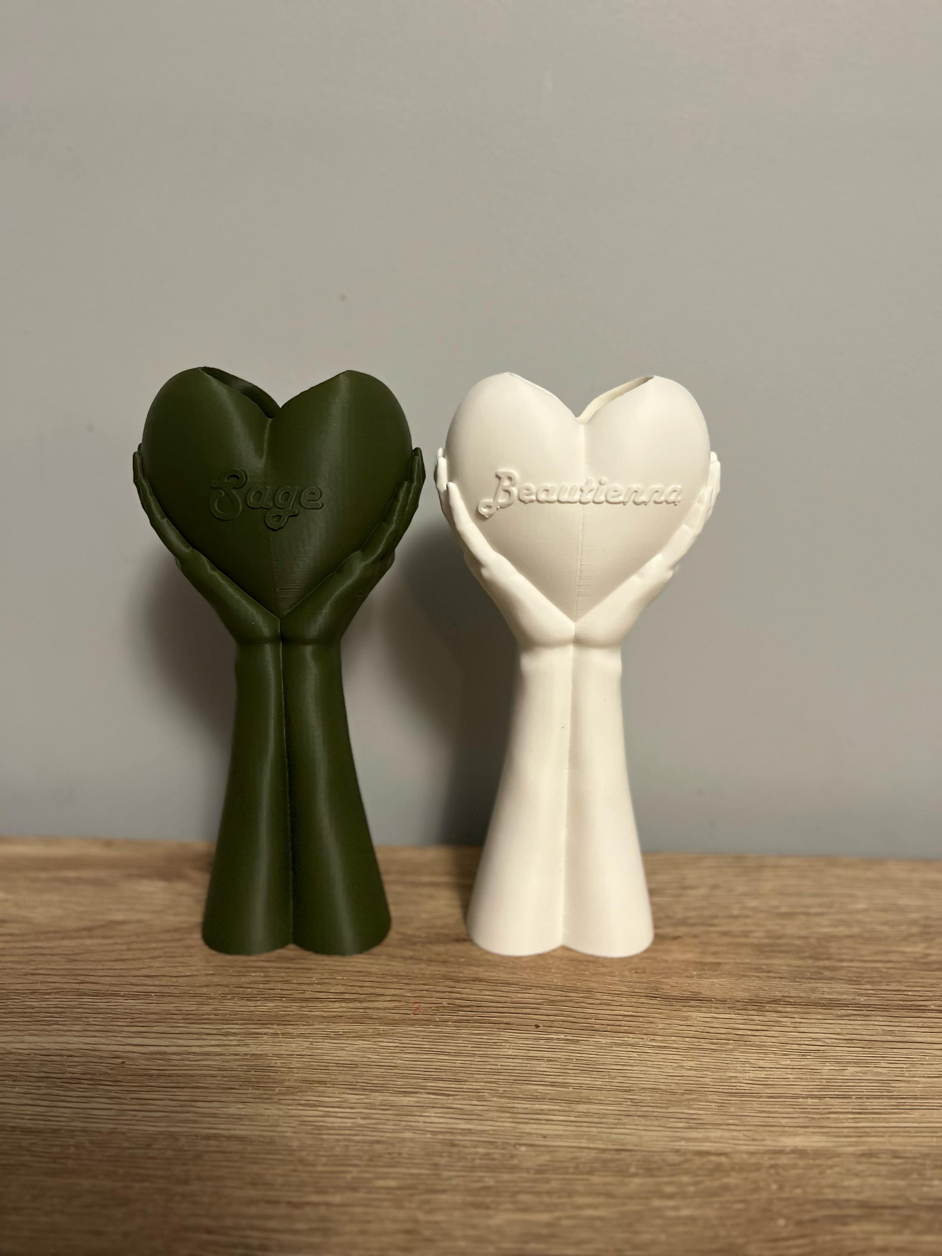 Two 3D-printed ‘Heart in Hands’ statues—one in sage green with the name 'Sage' and one in white, displayed on a wooden surface with a neutral wall background.