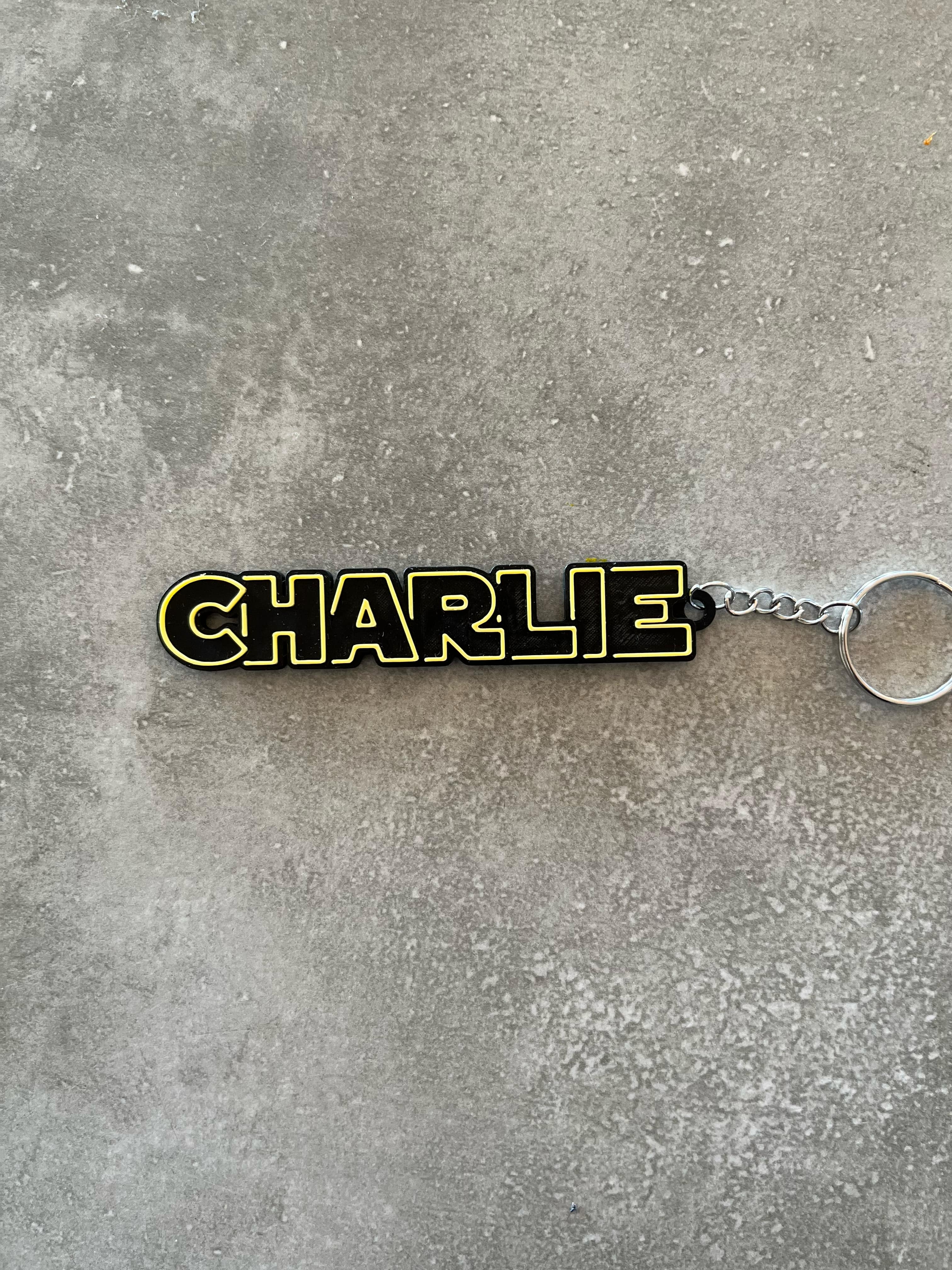 D-printed black keychain with the name ‘CHARLIE’ outlined in yellow, attached to a metal keyring and placed on a concrete surface.