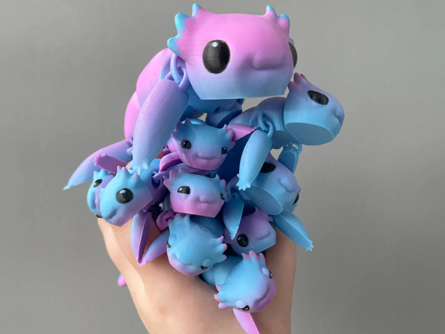 A hand holding a stack of tiny blue and pink 3D printed axolotl dragon toys against a gray background.