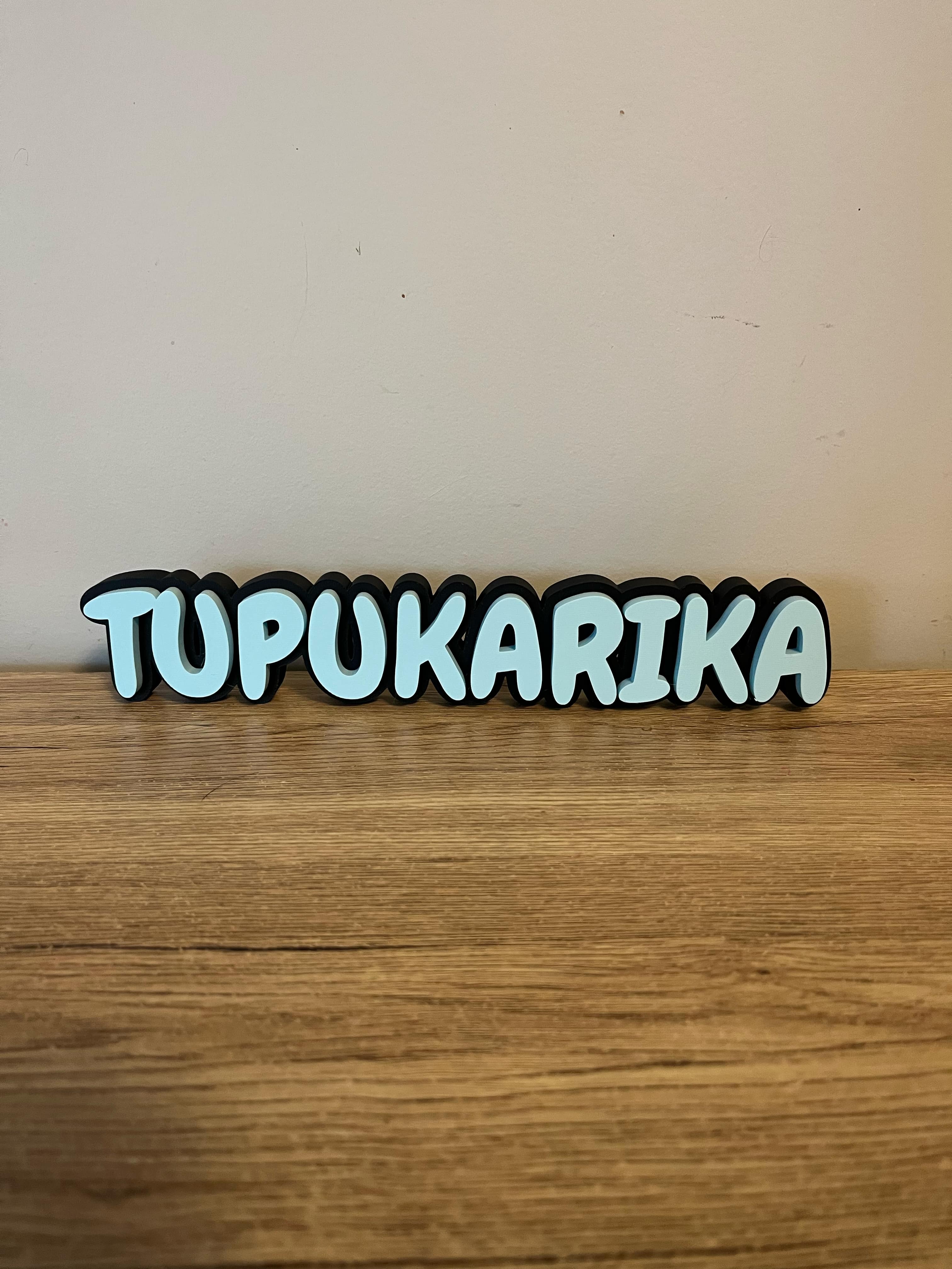 3D-printed name sign reading ‘TUPUKARIKA’ in light blue with a black outline, placed on a wooden surface with a soft beige wall in the background.