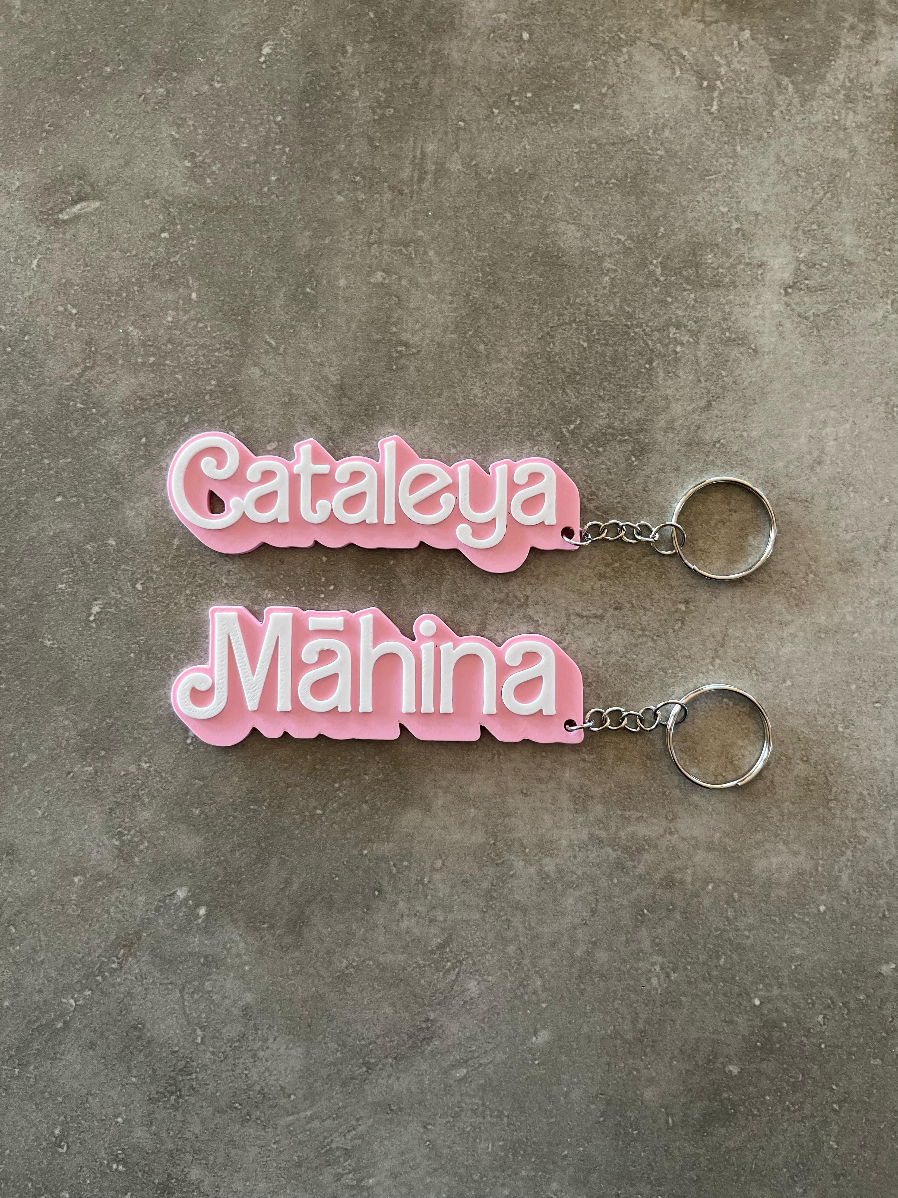 Two pink 3D-printed name keychains reading 'Cataleya' and 'Māhina' in white text, displayed on a grey concrete surface with attached metal keyrings.