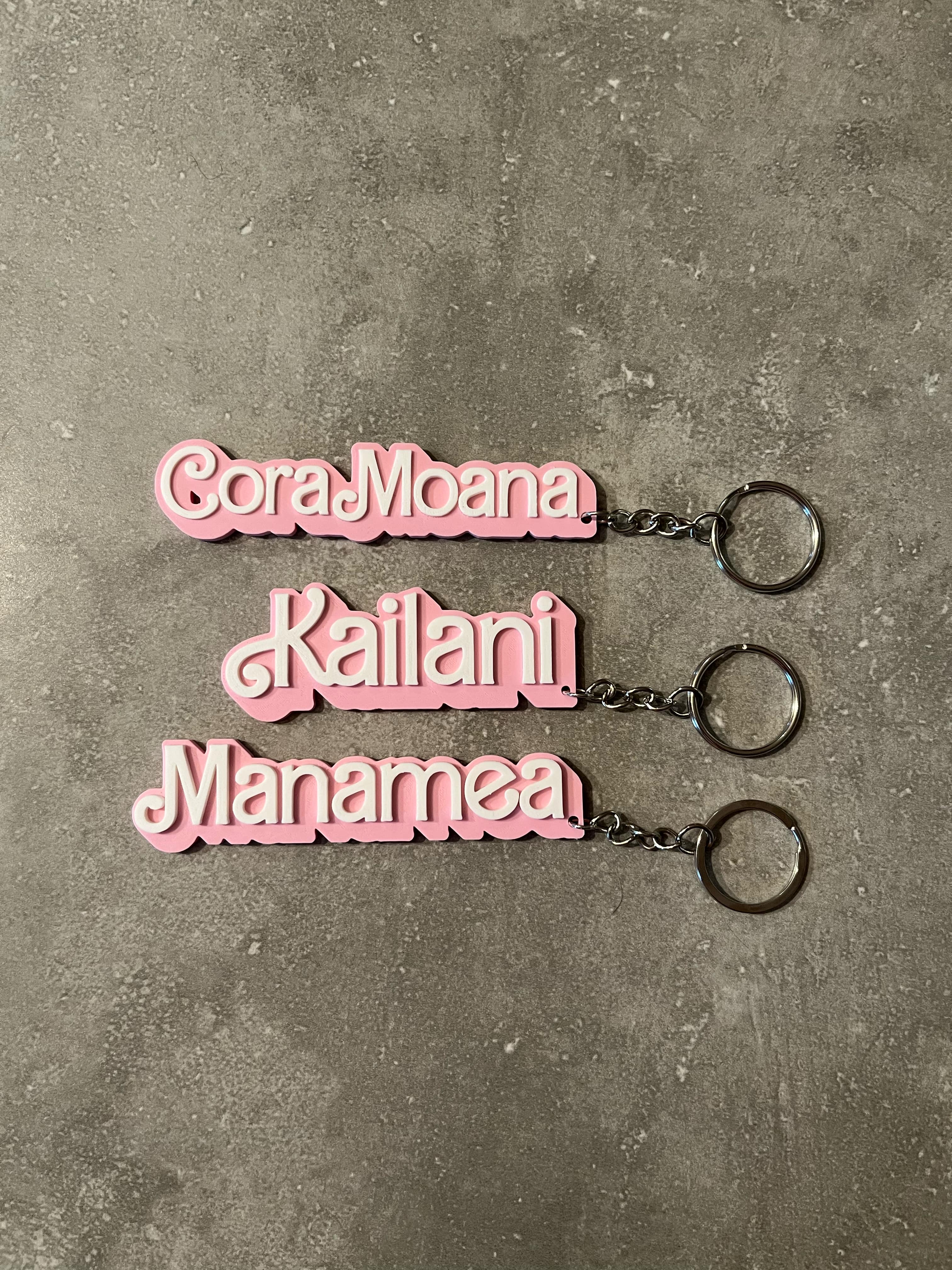 Three pink 3D-printed keychains with white text featuring the names 'Cora-Moana', 'Kailani', and 'Manamea', displayed on a grey concrete surface with attached metal keyrings.