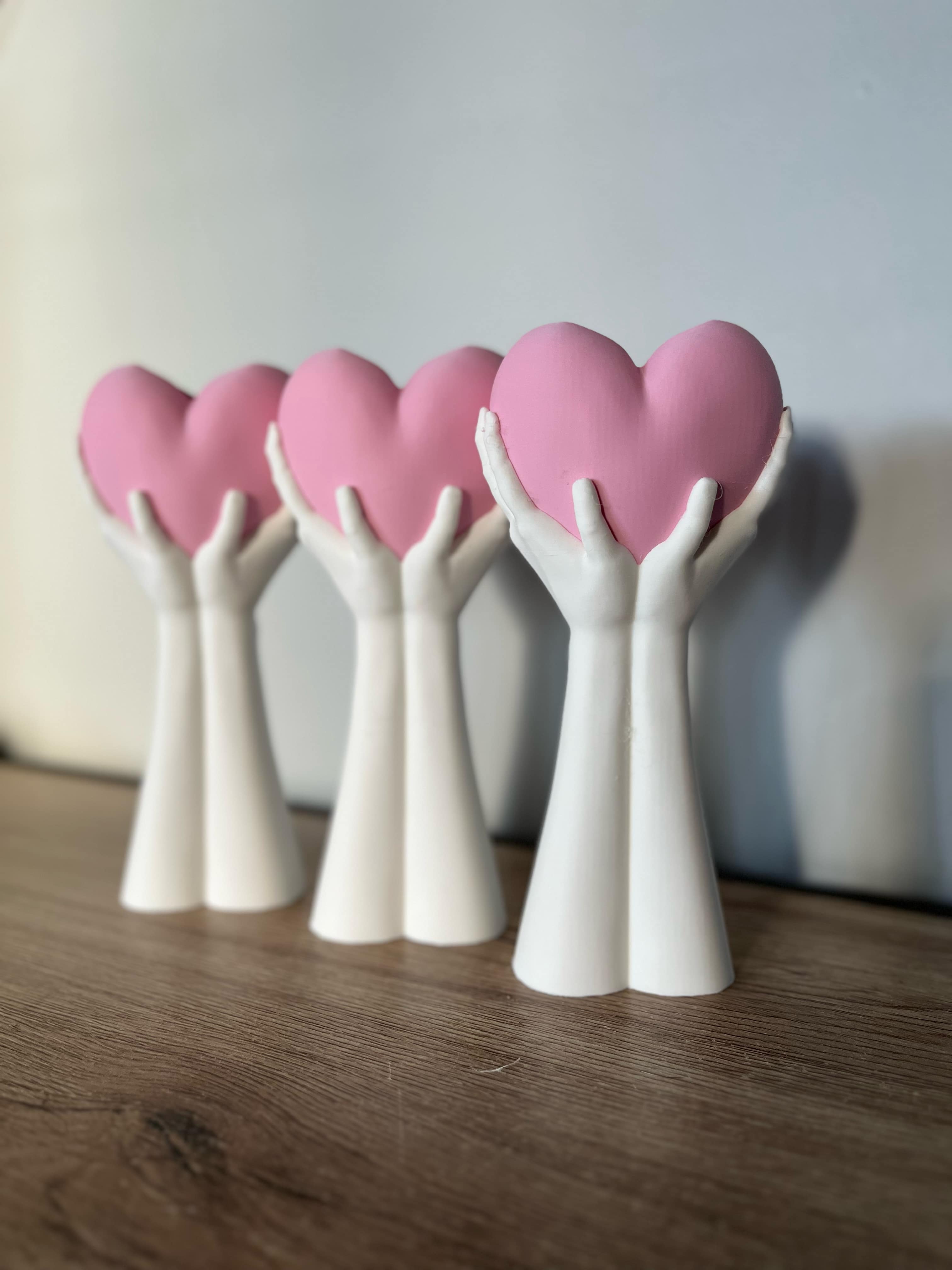 Five white 3D-printed hand sculptures holding pink hearts, arranged on a wooden surface with a soft grey background.