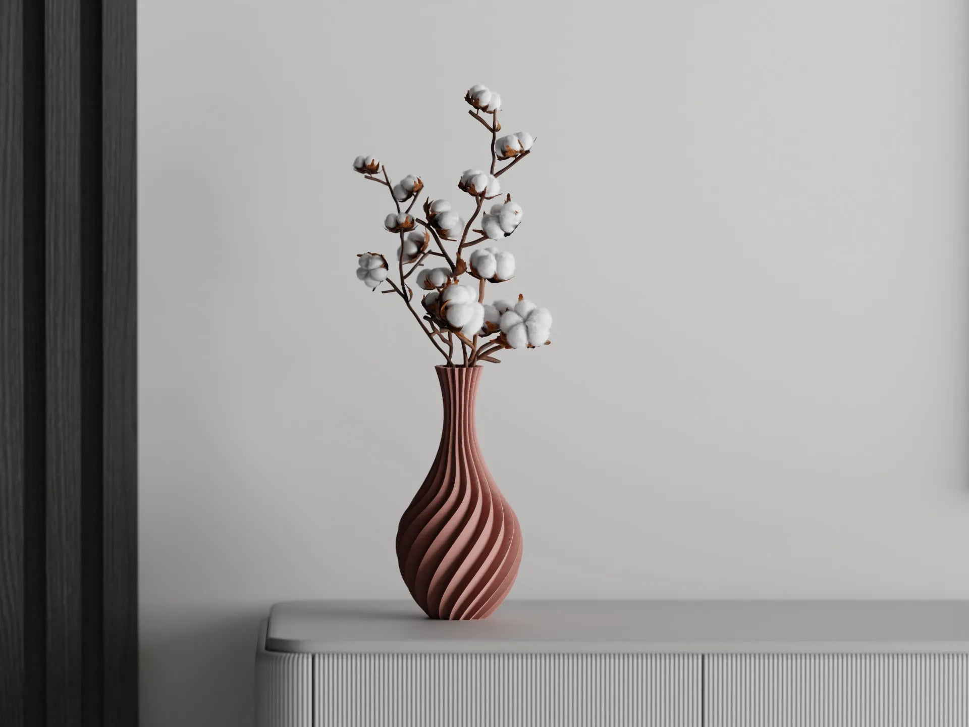 Curved terracotta 3D printed vase with spiral design, holding cotton branches, displayed on a light sideboard against a grey wall