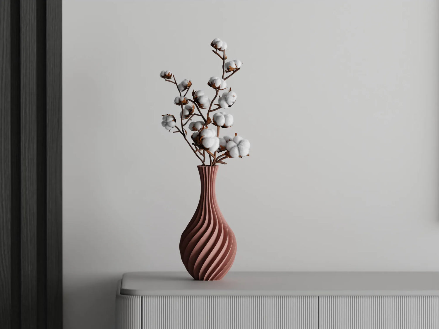 Curved terracotta 3D printed vase with spiral design, holding cotton branches, displayed on a light sideboard against a grey wall