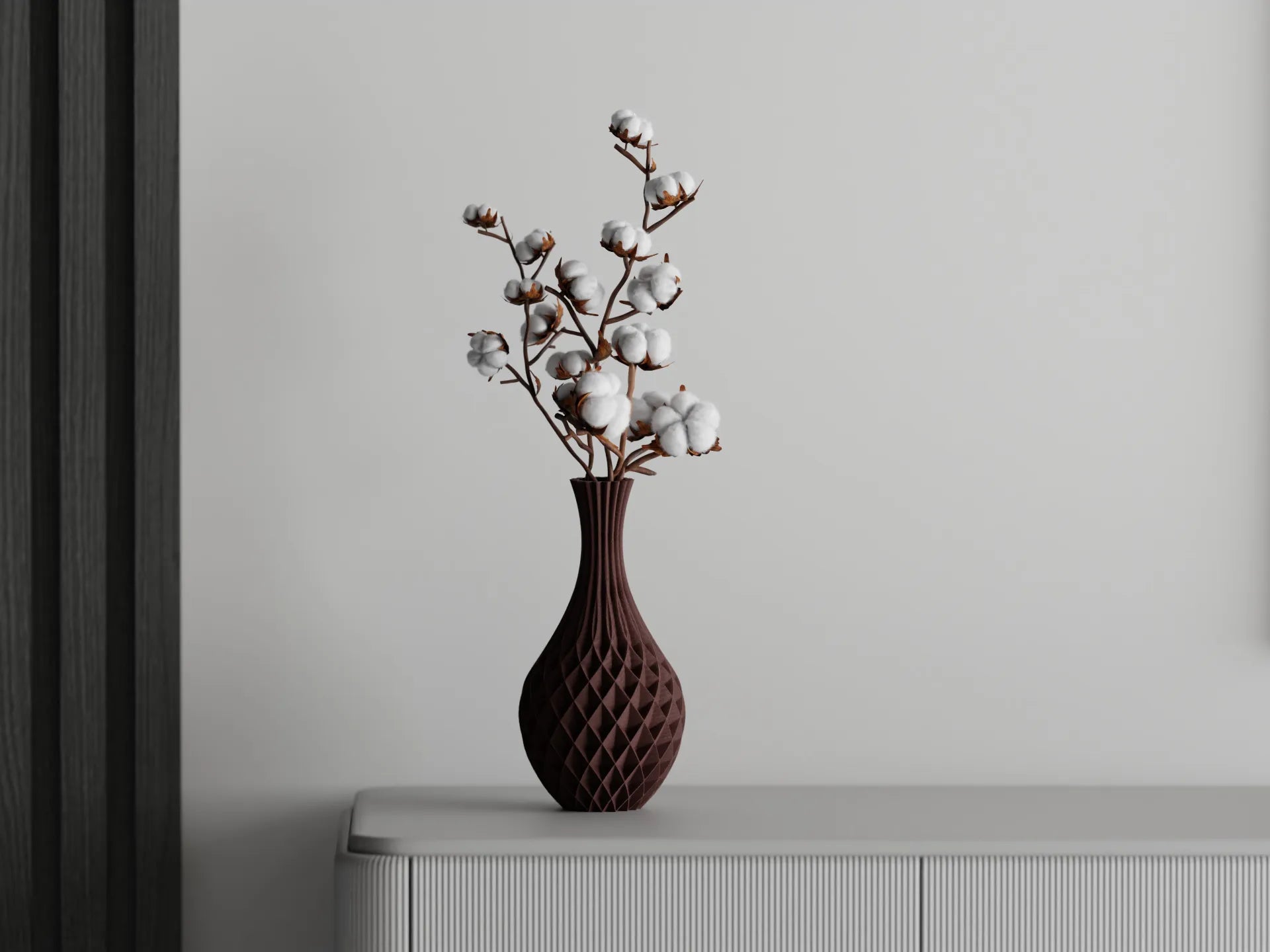 Tall maroon 3D printed vase with a faceted design holding cotton branches, placed on a white sideboard against a grey wall