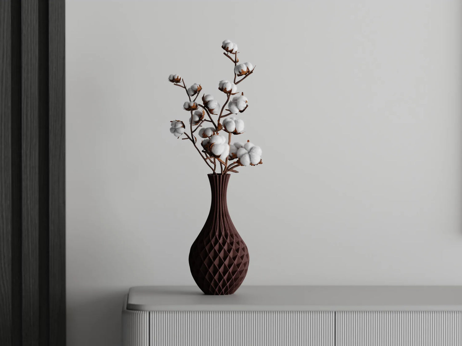 Tall maroon 3D printed vase with a faceted design holding cotton branches, placed on a white sideboard against a grey wall
